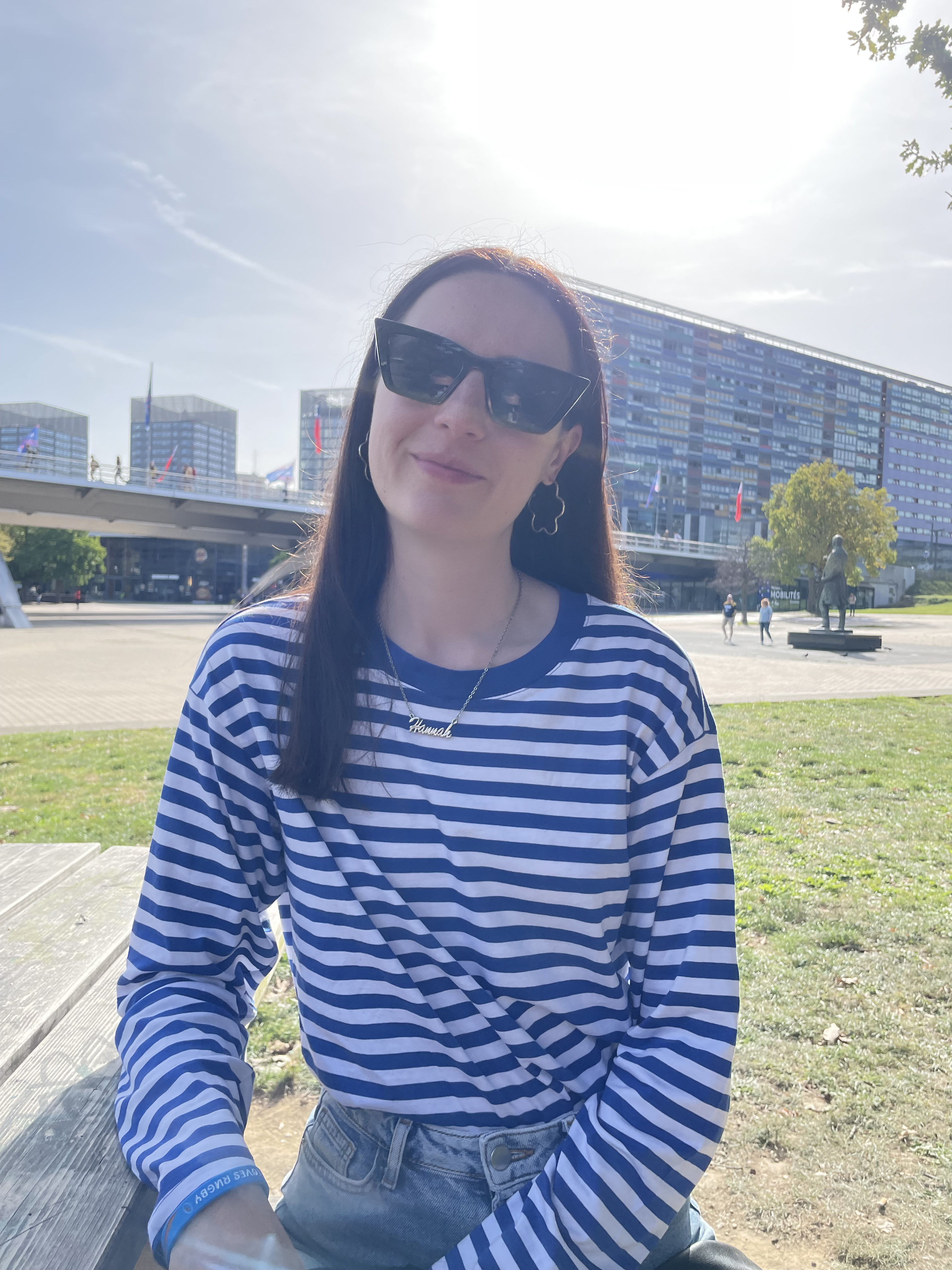 Hannah sitting in Lille, France wearing a blue and white striped shirt and black sunglasses, smiling at the camera
